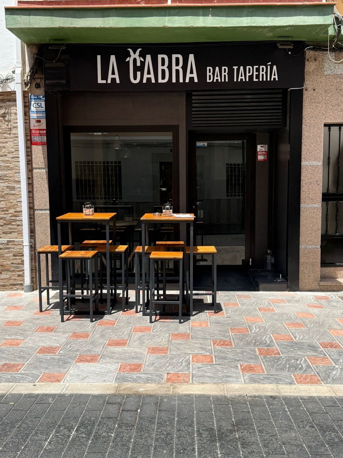The exterior of La Cabra - Bar Tapería features a modern entrance with high tables and chairs outside.