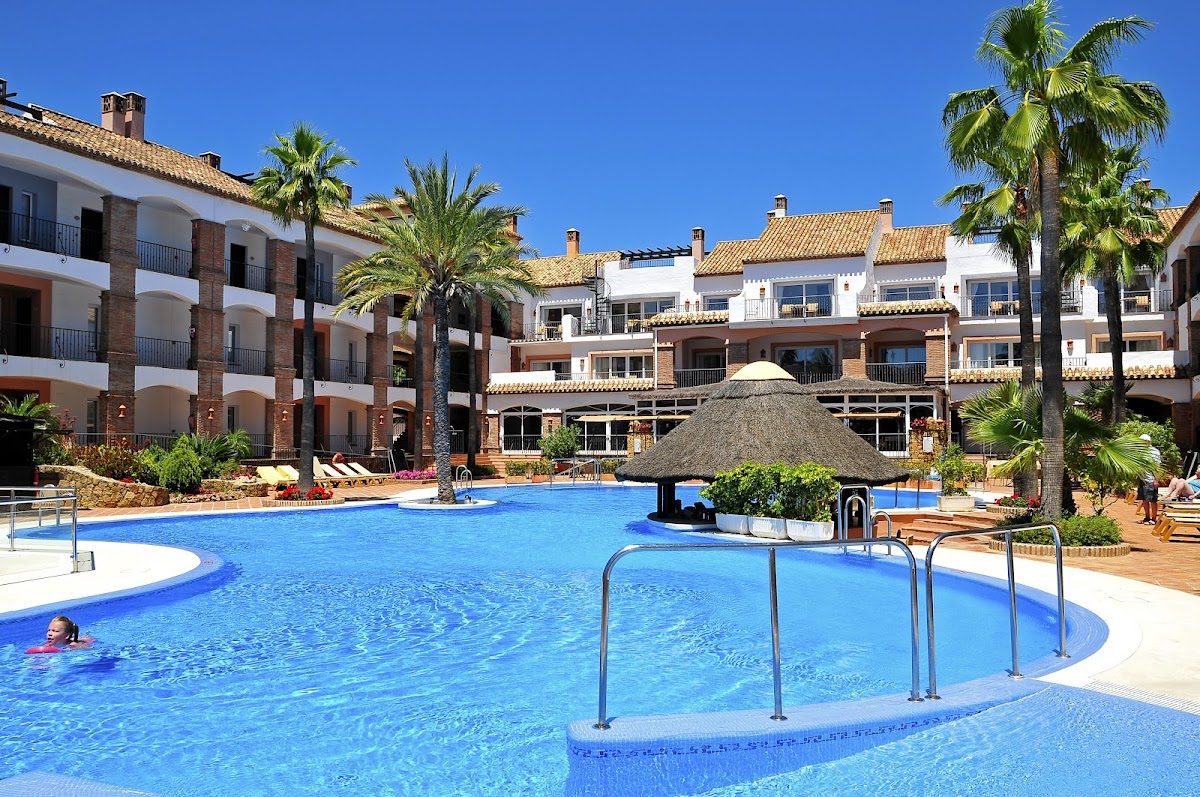 La Cala Hotel features a large outdoor pool surrounded by palm trees and a thatched-roof bar.