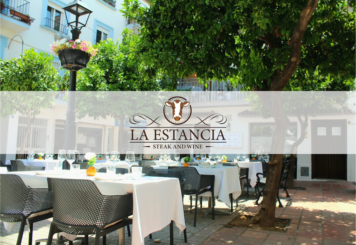 El restaurante La Estancia Steak and Wine cuenta con una terraza al aire libre con mesas dispuestas bajo los árboles en una plaza.