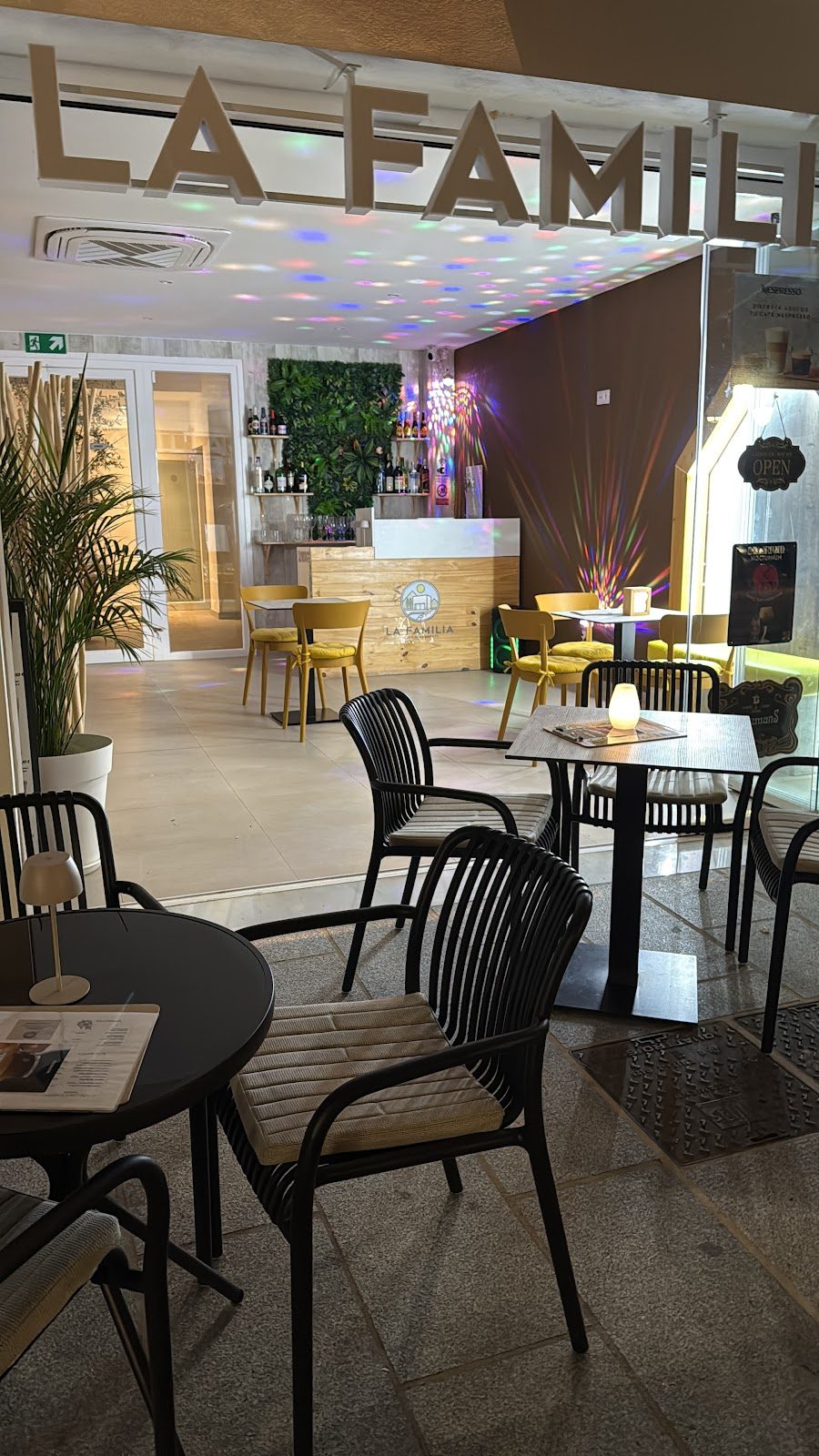 El bar La Familia Mijas presenta un interior moderno con asientos, una barra y efectos de iluminación coloridos.