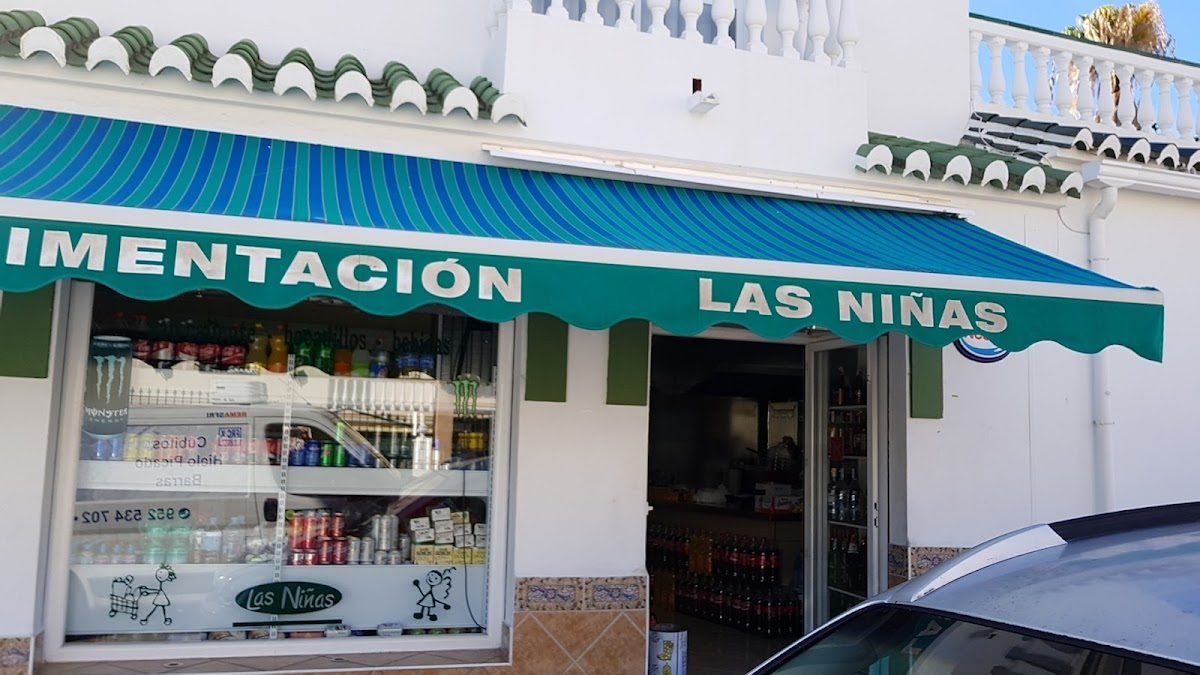 Exterior view of Las Niñas shop with a green and blue striped awning and a display of beverages inside.