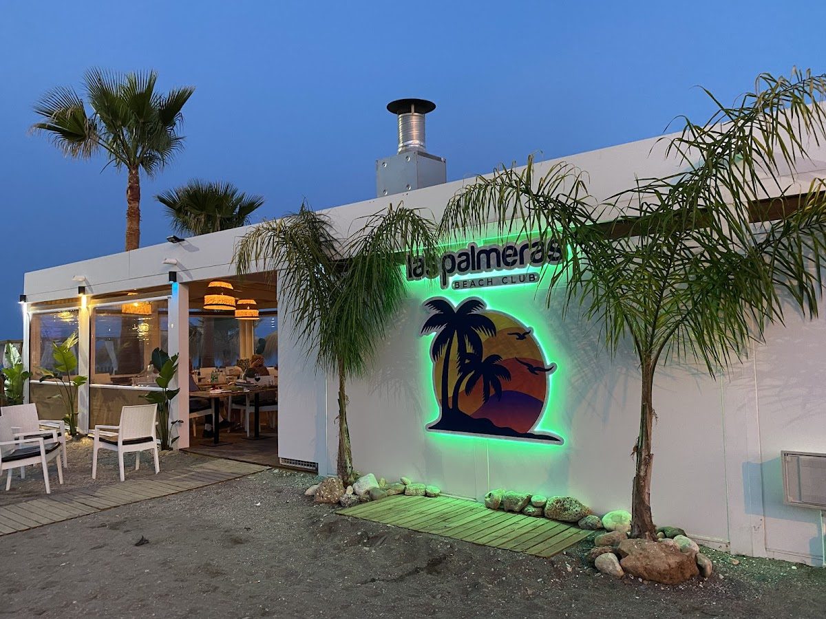 The exterior of Las Palmeras Beach Club features a neon sign, palm trees, and a sandy beach setting.