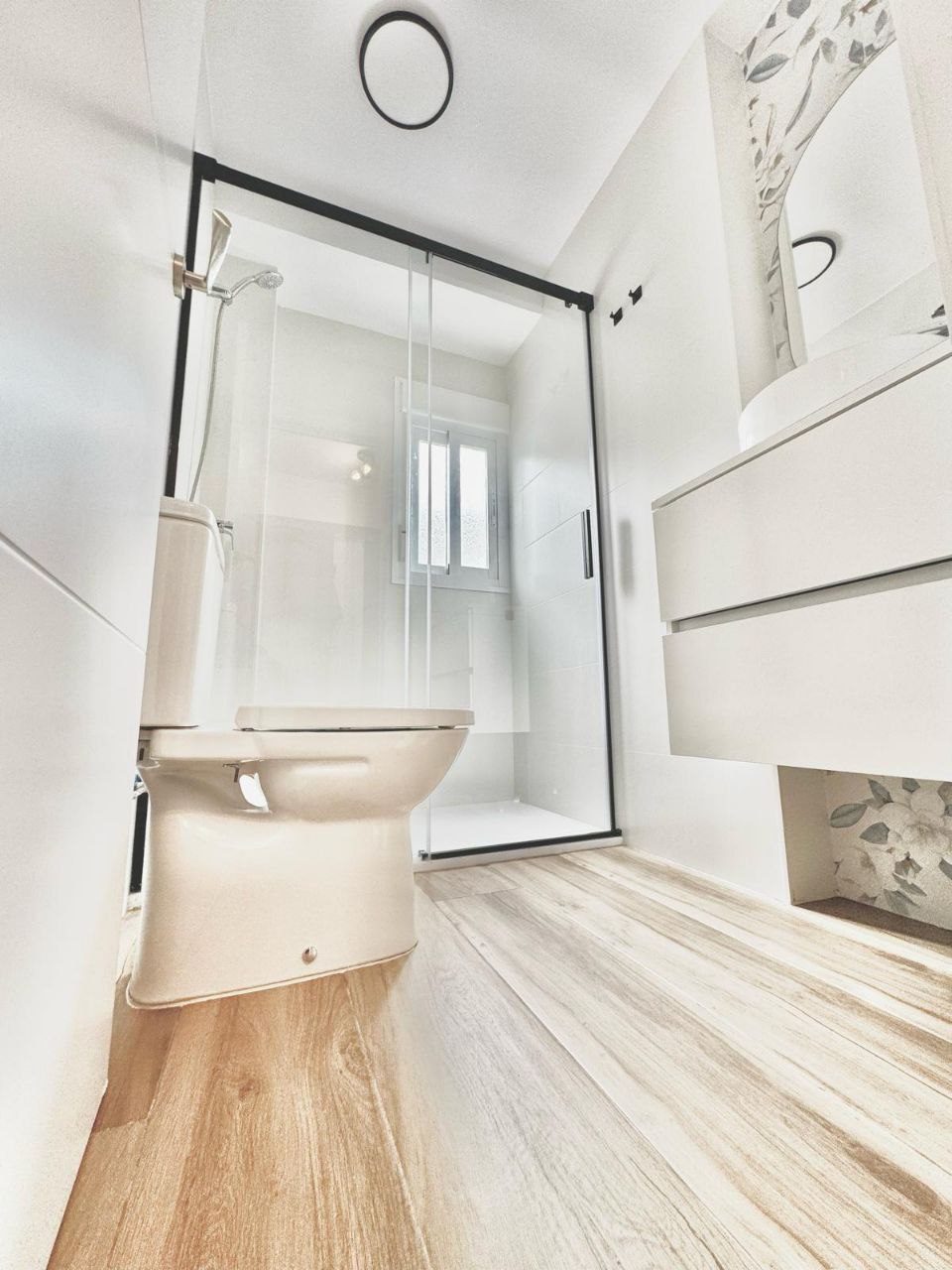 Baño moderno con ducha de cristal, grifería blanca y suelo de madera en Las Rejas Reformas Torremolinos.