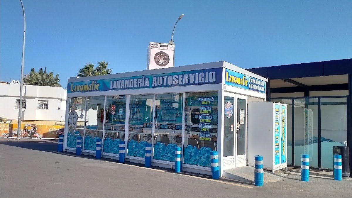 Vista exterior de Lavandería Autoservicio Madese - Lavomatic con letreros azules y blancos y máquinas de lavandería visibles en el interior.