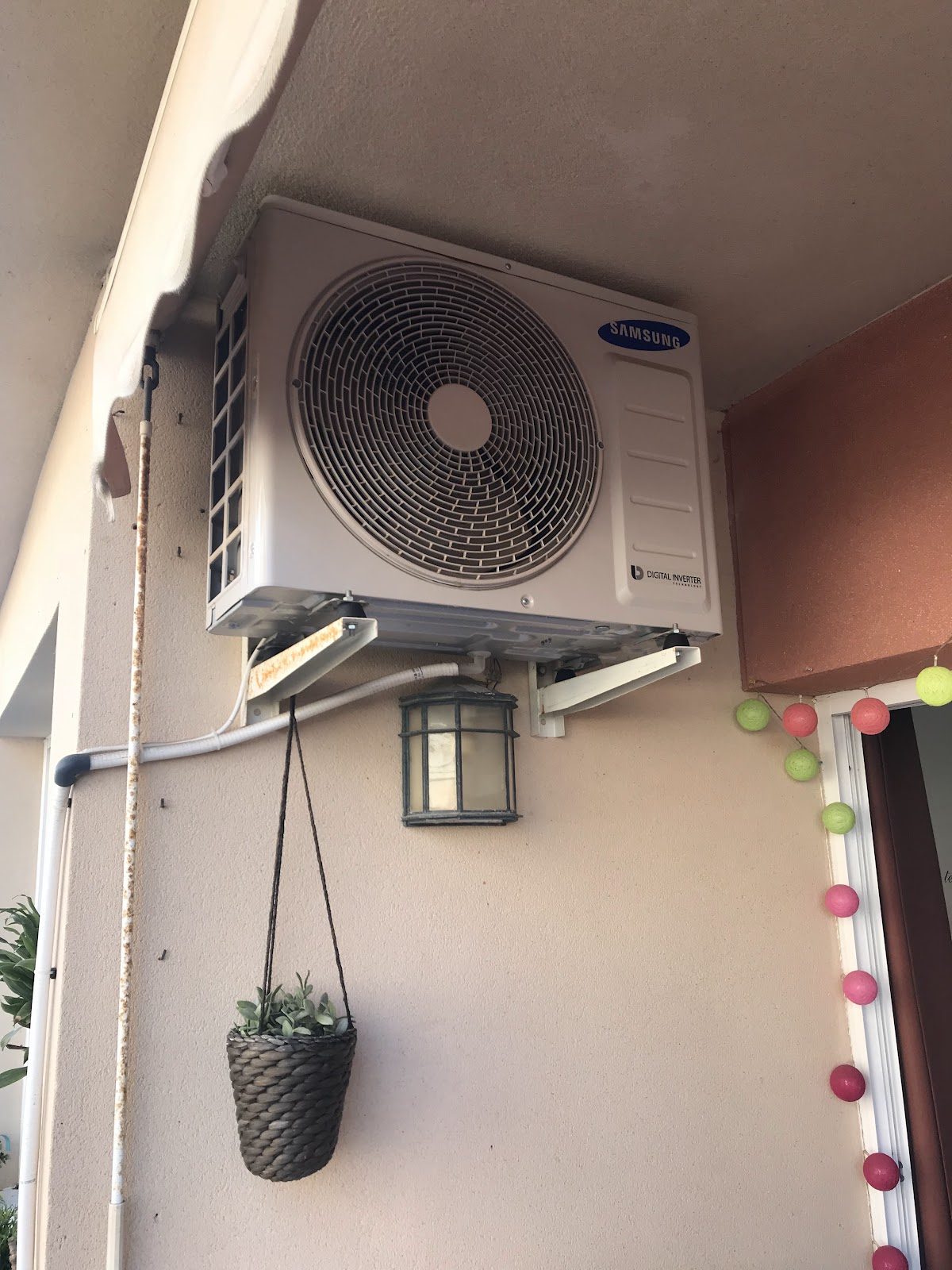 An air conditioning unit mounted on a wall, with a hanging plant and decorative lights nearby.