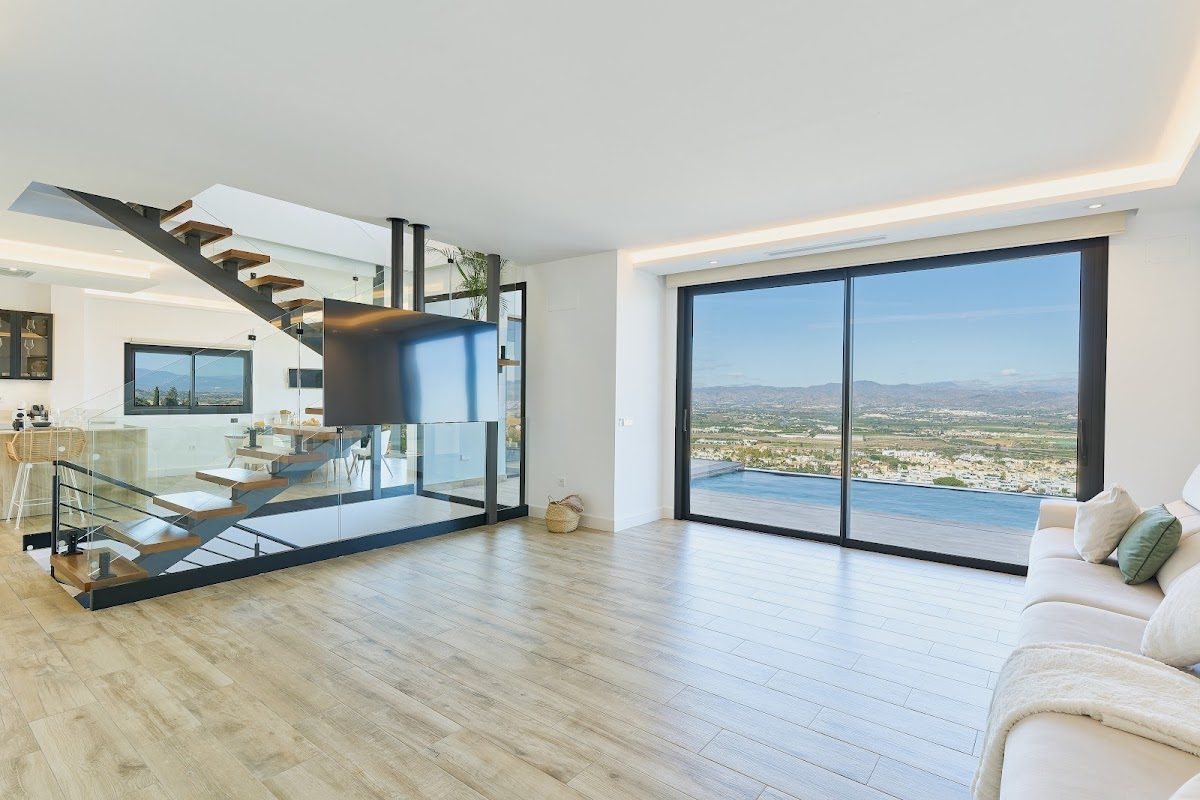 Interior moderno de un luminoso salón con grandes ventanales que ofrecen vistas panorámicas y una elegante escalera.