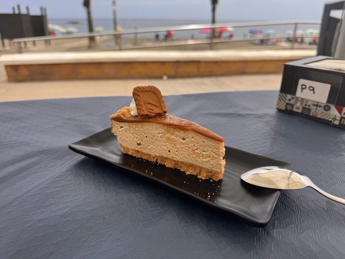 Una porción de postre en un plato negro con una cuchara, con el paisaje playero de Macai de fondo.