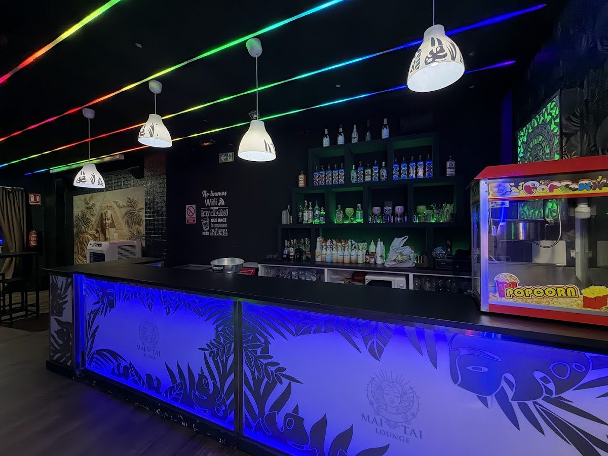 The MAI-TAI lounge features a vibrant bar with colorful lighting, shelves of drinks, and a popcorn machine.