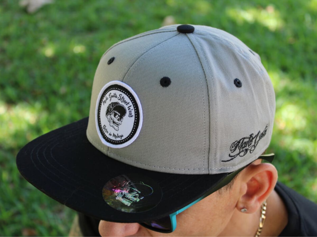 Primer plano de una gorra snapback gris y negra con el logotipo de Mala Guita Streetwear.
