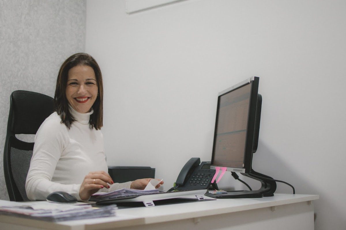 A smiling woman sits at a desk with a computer and phone in M&C Consulting's office.