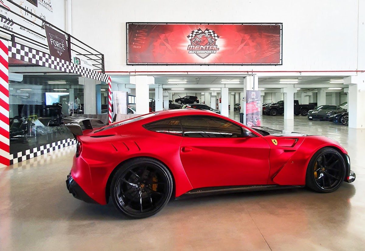 Un elegante coche deportivo rojo expuesto en el interior de la sala de exposiciones de Marbella Motorsports, con otros vehículos visibles al fondo.