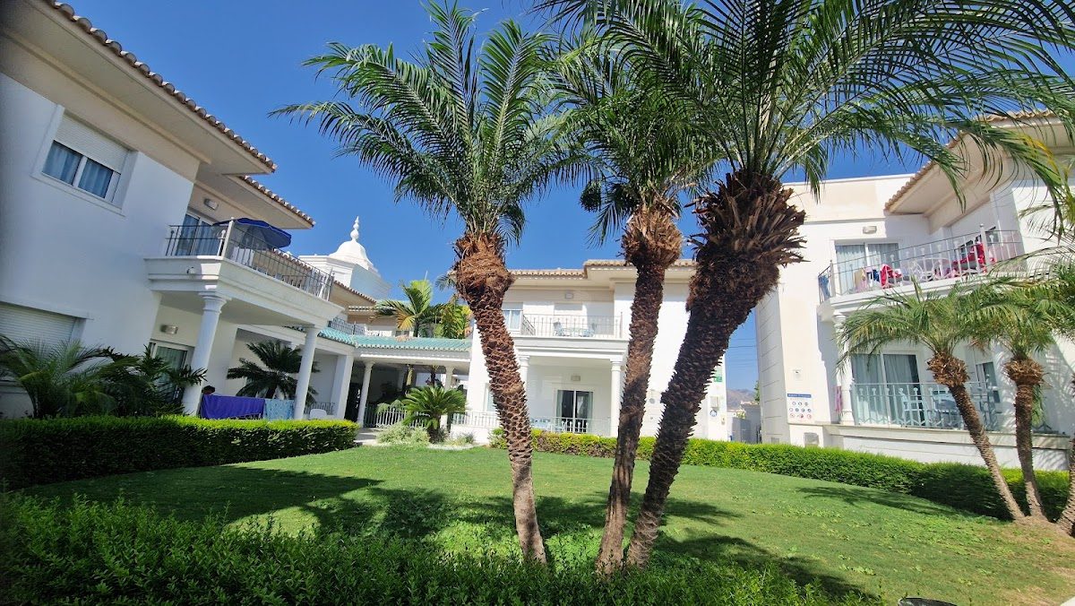 Marina Turquesa Aparthotel features a lush garden with palm trees and well-maintained white buildings.