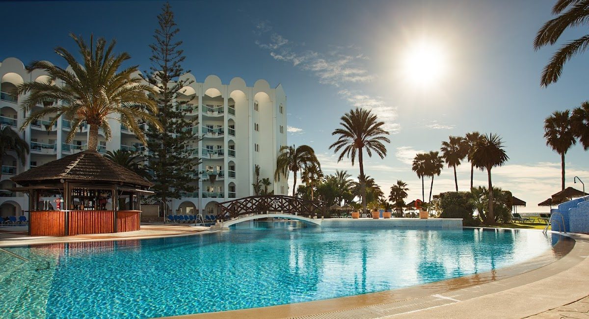 The image shows a large outdoor pool surrounded by palm trees and a white hotel building at Marinas de Nerja Beach & Spa Aparthotel.