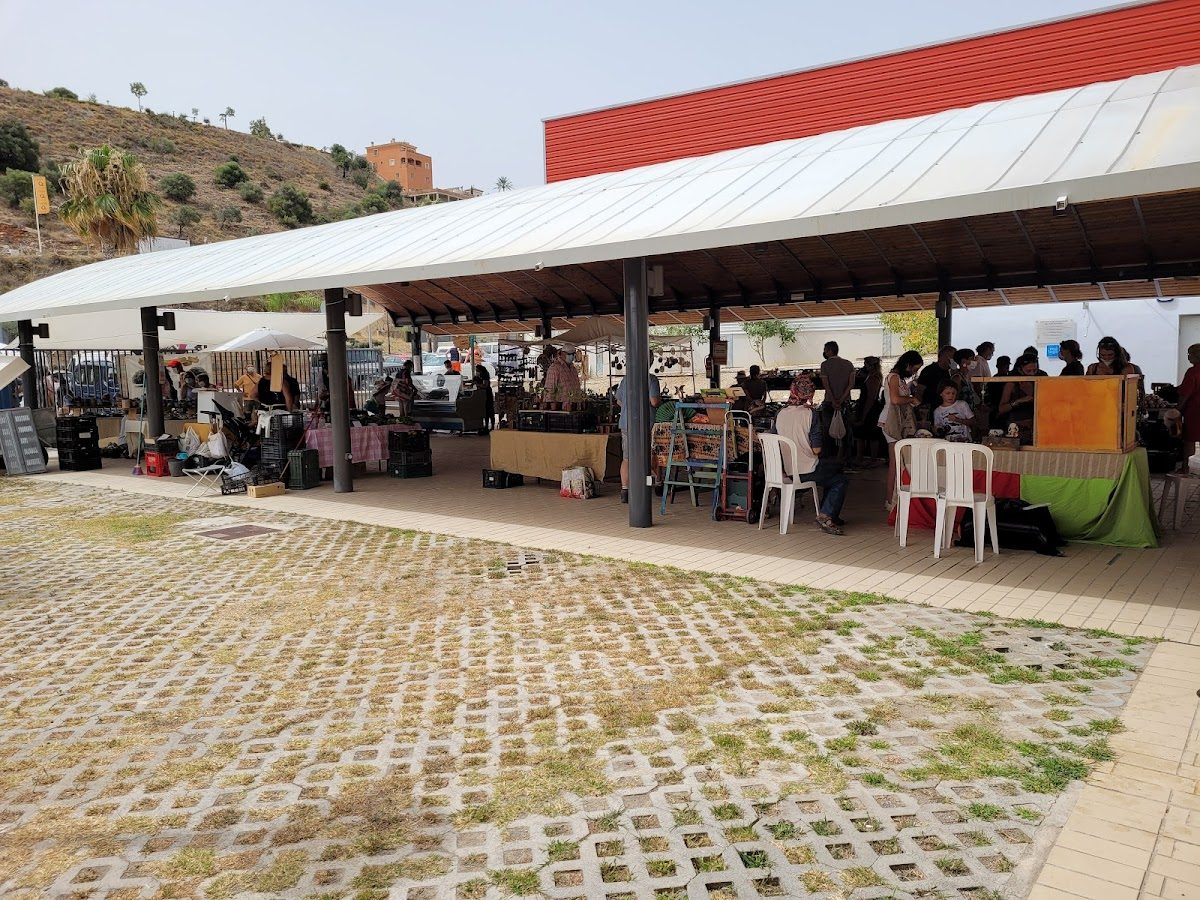 Mercado Agroalimentario de Coín: Mercado al aire libre con diversos puestos, gente haciendo sus compras y un pintoresco paisaje de ladera como telón de fondo.