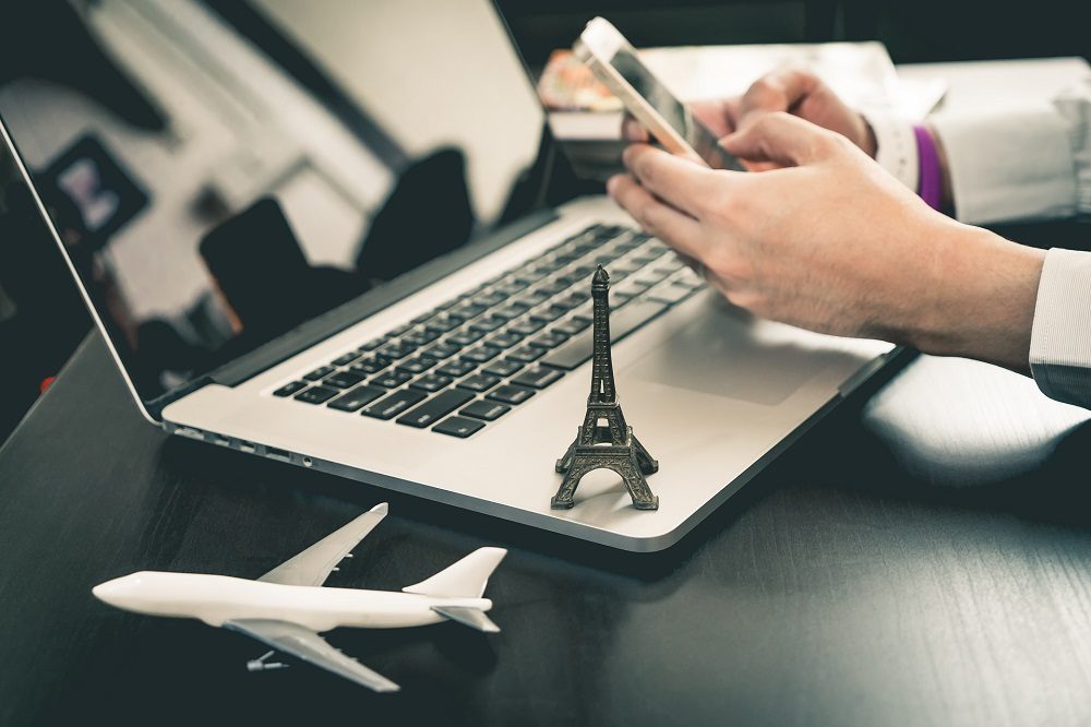 Una persona usando un teléfono inteligente y una computadora portátil con una réplica en miniatura de la Torre Eiffel y un avión sobre el escritorio.