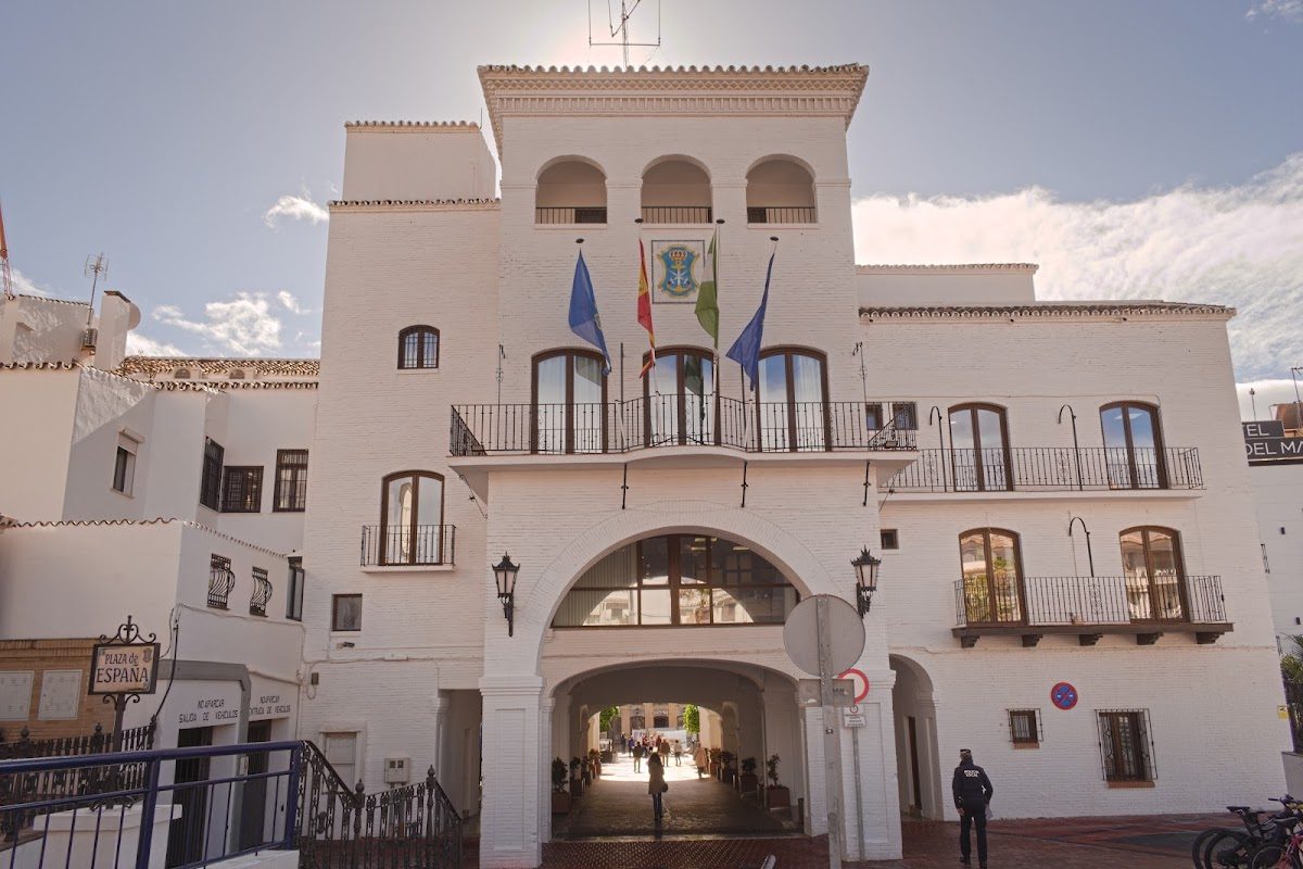 The Nerja Tourist Office features a white building with arches, balconies, and flags in a sunny plaza.