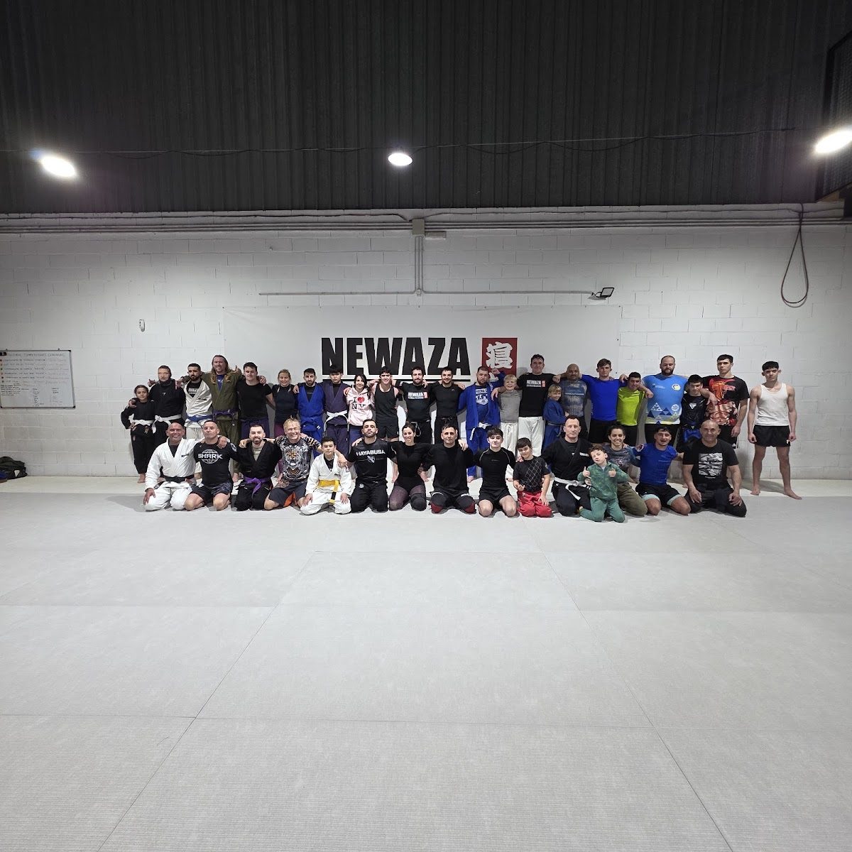 A large group of people in training gear poses on a mat inside Newaza Jiujitsu Academy.