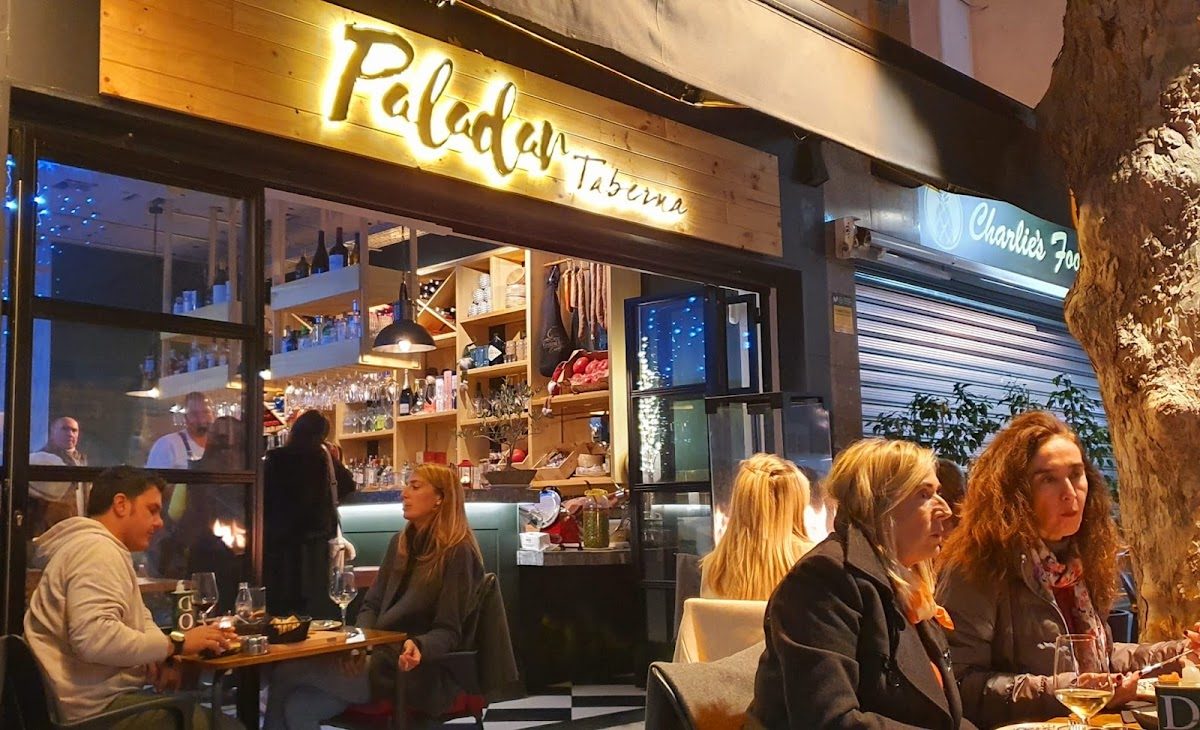 Paladar Taberna cuenta con una acogedora zona de comedor al aire libre donde los clientes disfrutan de sus comidas y un interior elegante que se puede apreciar a través de las ventanas.