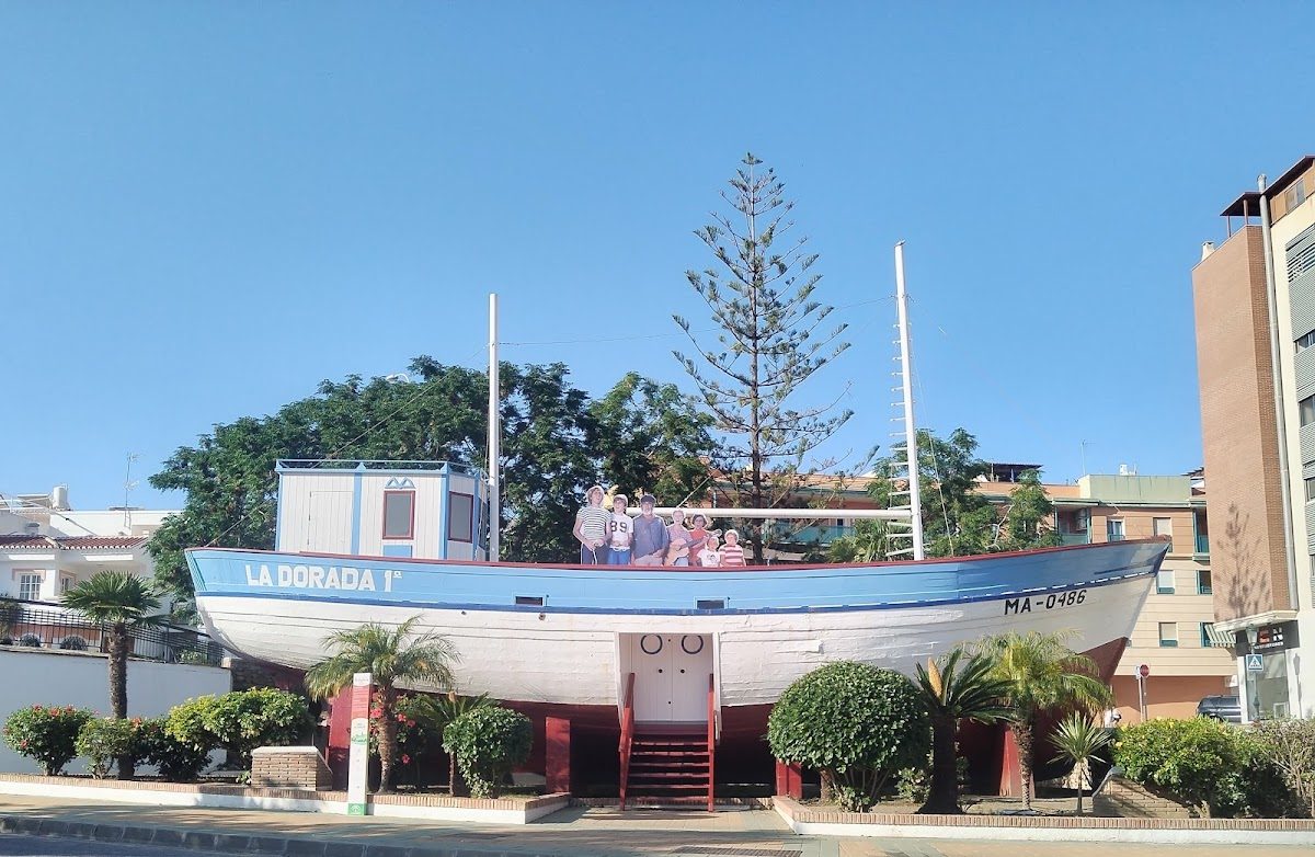 Una gran escultura de un barco llamada "La Dorada I" con personas posando delante, rodeada de vegetación en el Parque Verano Azul.