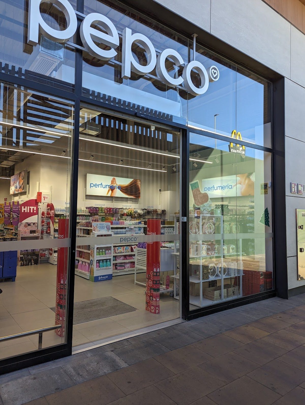 La tienda Pepco cuenta con una entrada de cristal con expositores de productos de belleza y un McDonald's en las inmediaciones.