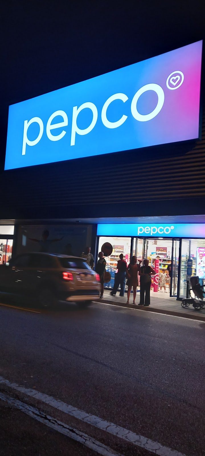 Exterior de una tienda Pepco por la noche, con letreros llamativos y gente esperando afuera.