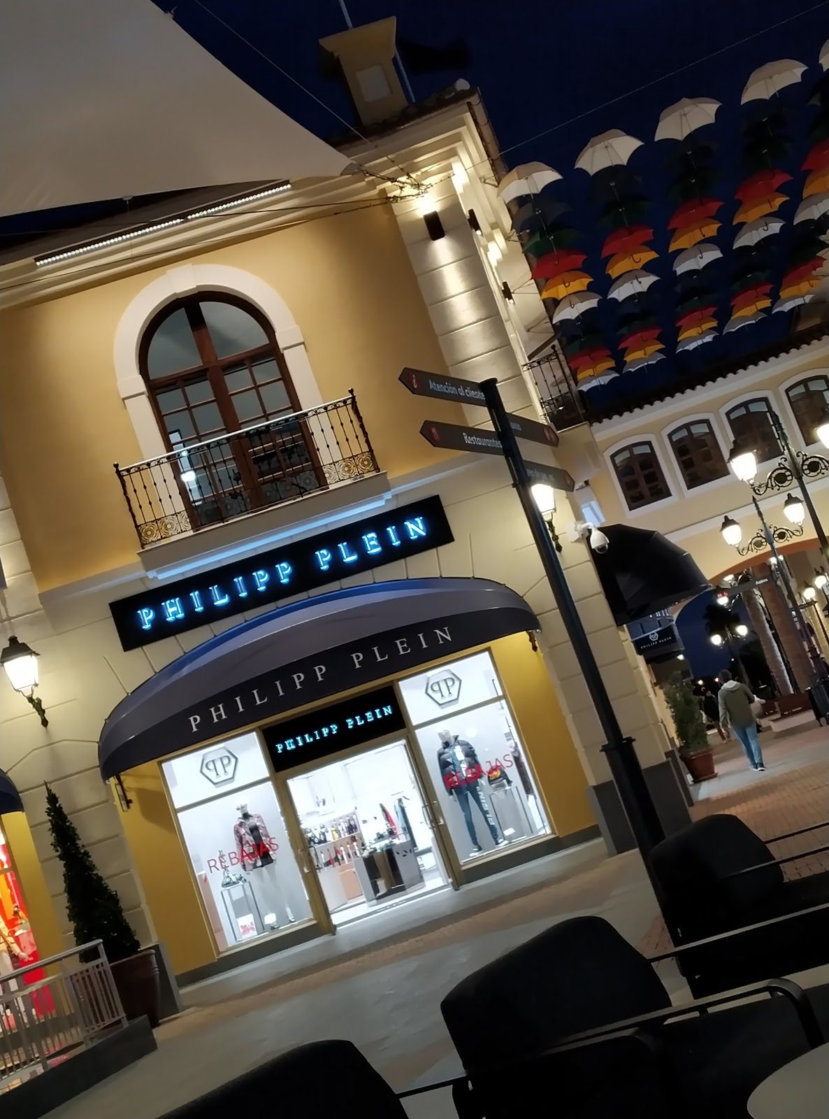 Fachada de la tienda Philipp Plein en Málaga, con letreros iluminados y coloridos paraguas en el techo.