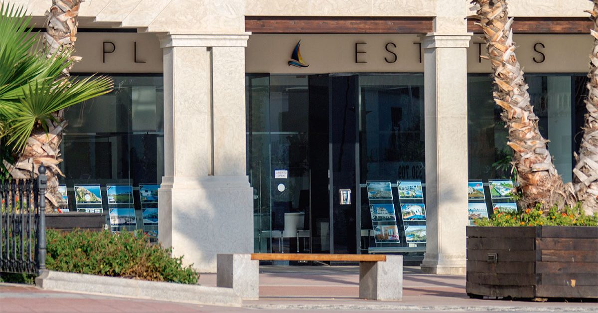 The entrance of Plaza Estates features large windows displaying property listings and palm trees nearby.
