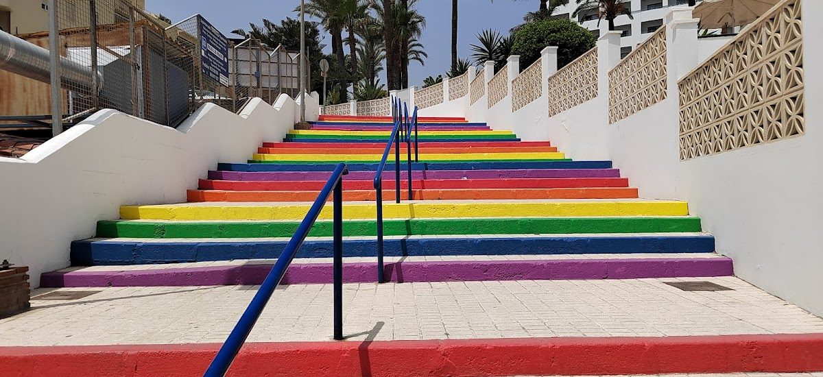 Escalones de colores del arcoíris que conducen a una escalera en Nerja, rodeada de palmeras y edificios.