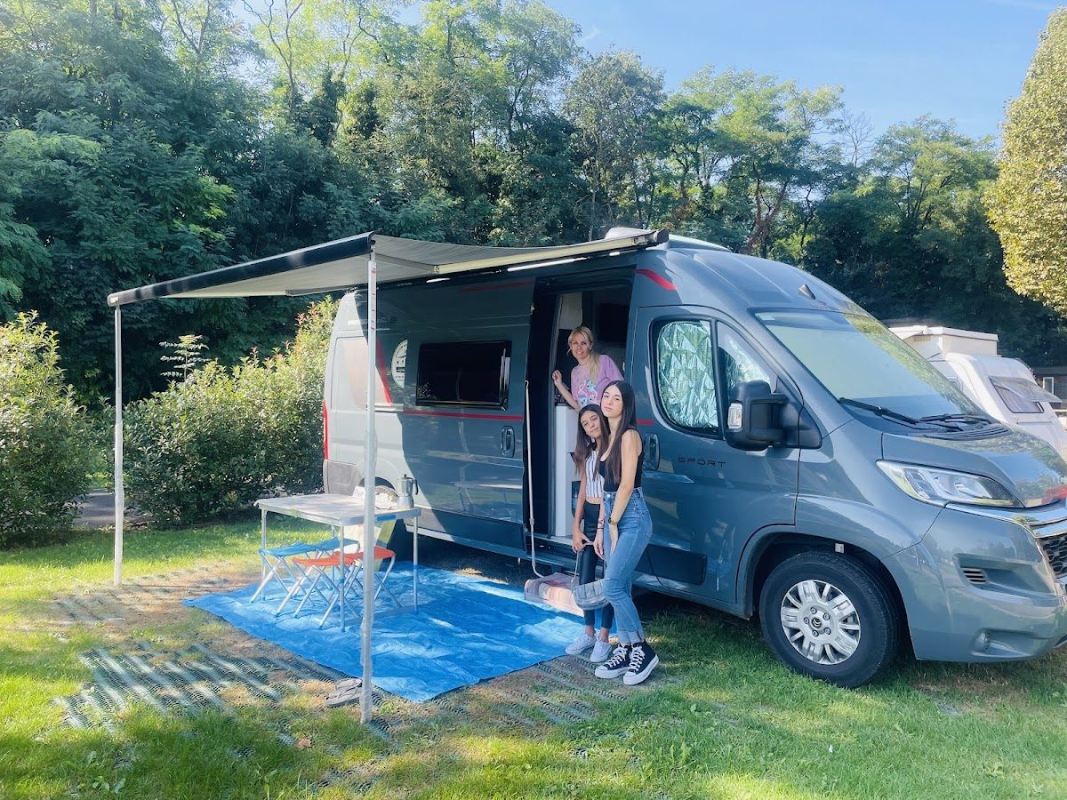 Una autocaravana gris con toldo aparcada sobre el césped, con tres personas de pie fuera.