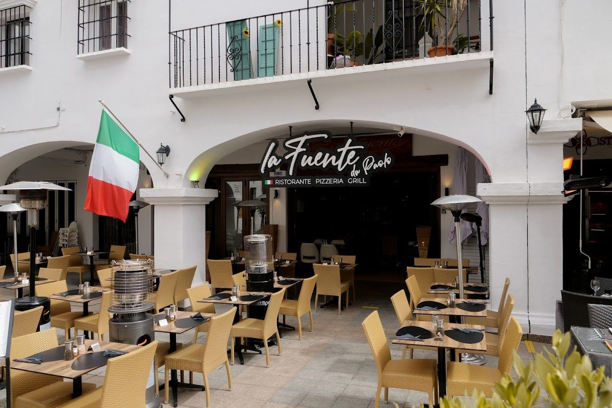 Outdoor seating area with tables and chairs, featuring an Italian flag and the "La Fuente" sign.