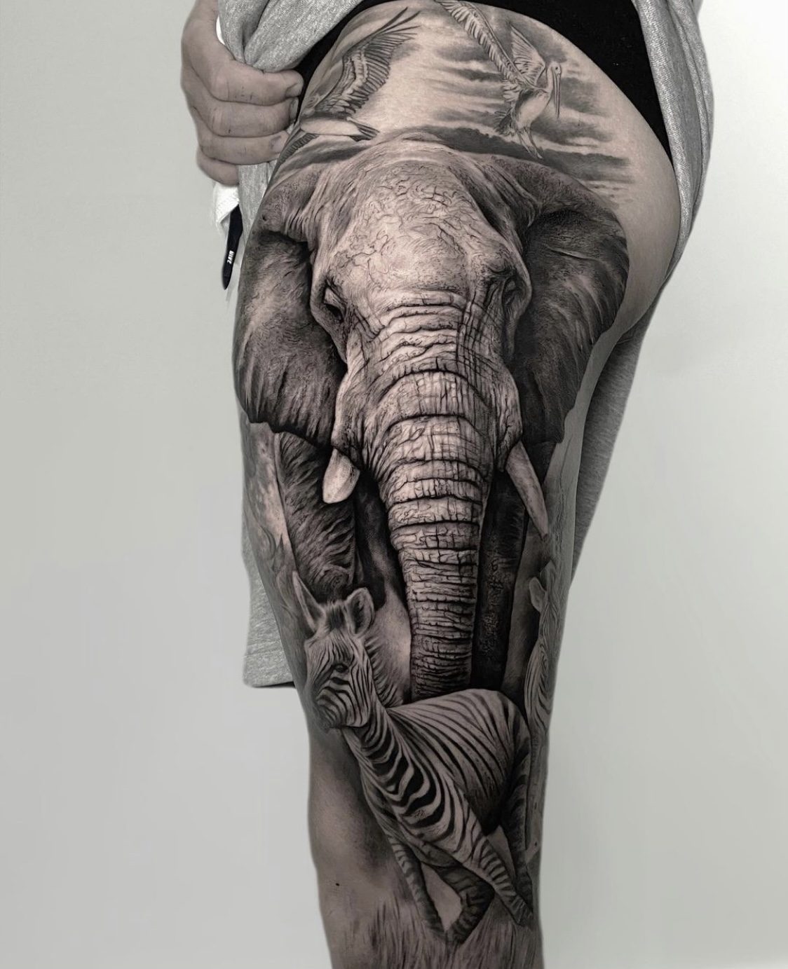 A detailed black and gray tattoo of an elephant and zebra on a thigh.
