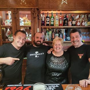 Cuatro empleados sonríen detrás de la barra del Styles Music Bar, con una variedad de botellas expuestas en los estantes.