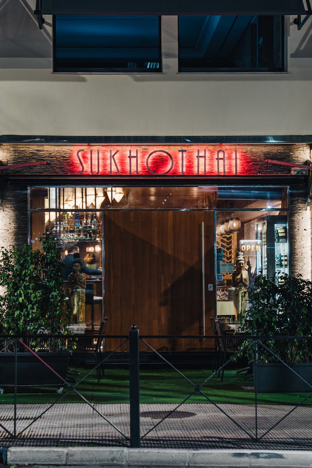 The entrance of Sukhothai by Thai Gallery features wooden doors, glowing red signage, and decorative plants.