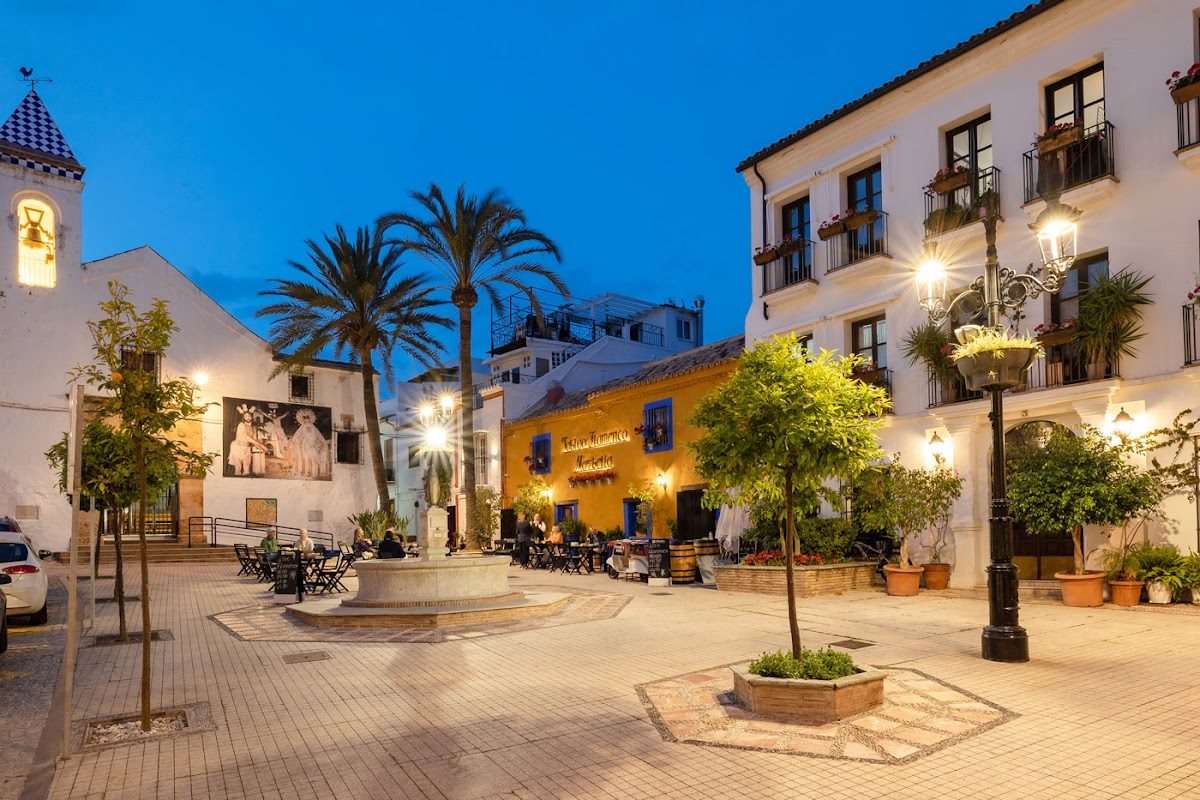Plaza con palmeras, una fuente y el vibrante exterior del Tablao Flamenco Marbella al atardecer.