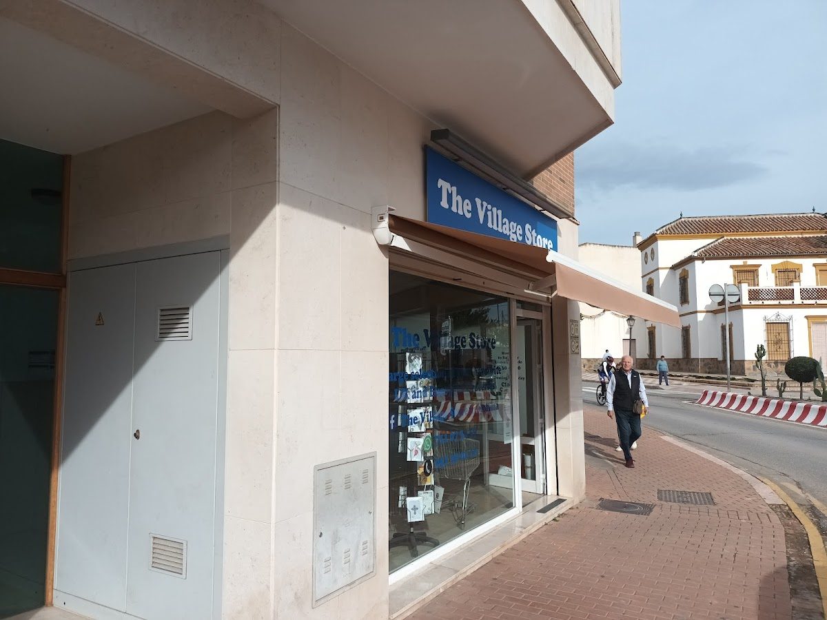 La tienda Village Store en Mollina tiene una fachada con letreros azules y un peatón que pasa caminando.