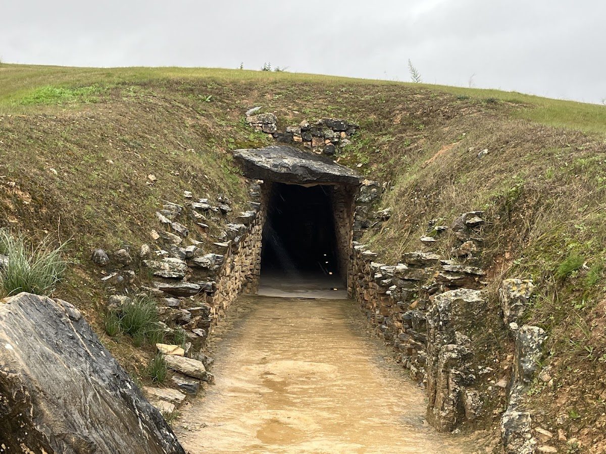 Tholos de El Romeral cuenta con una entrada de piedra que conduce a un montículo cubierto de hierba, rodeado de terreno rocoso.