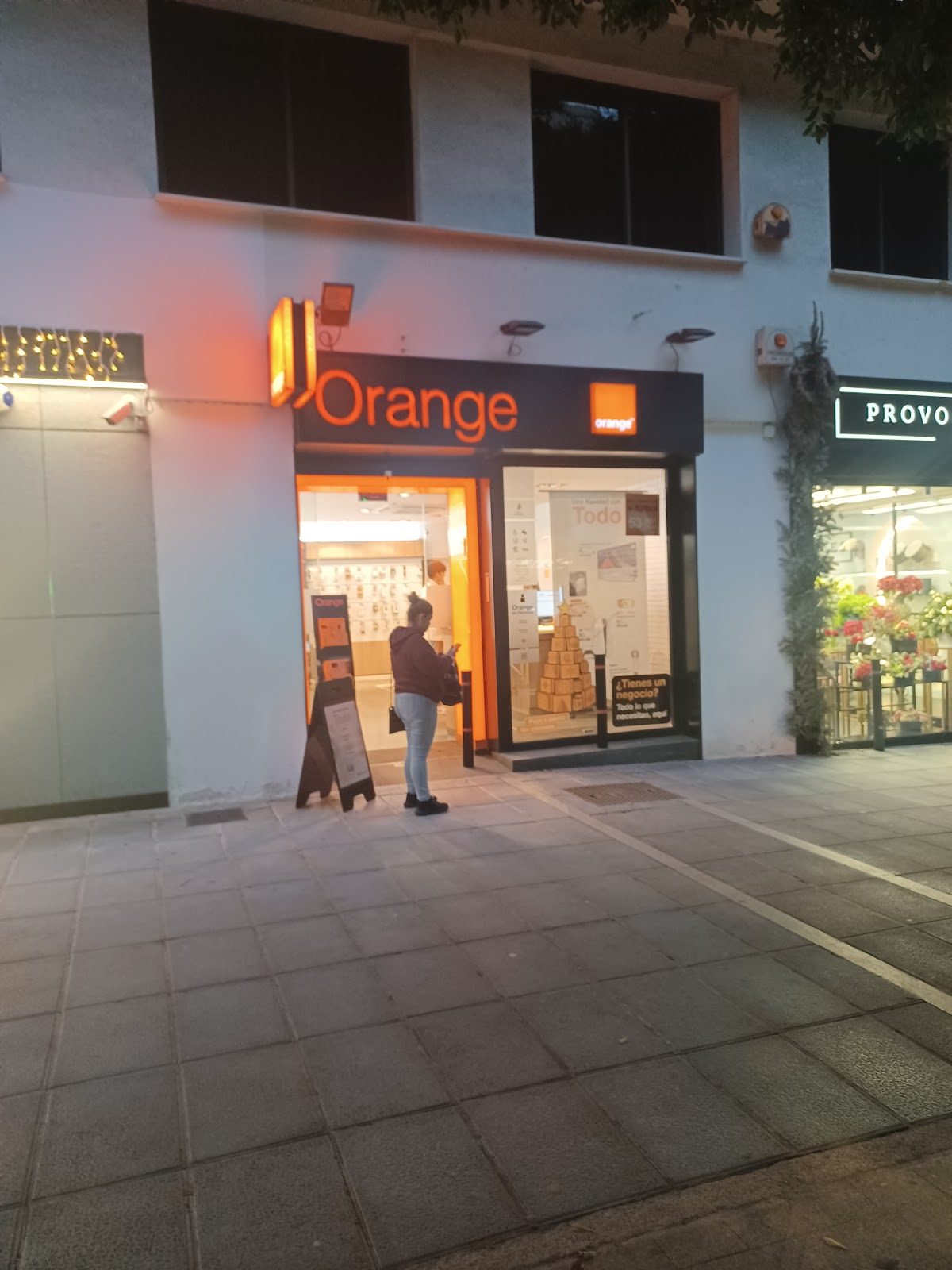 En el exterior de Tienda Orange se puede ver un letrero de color naranja brillante y a un cliente de pie afuera.
