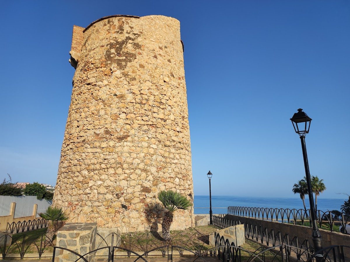 La Torre Vigía de El Cantal es una torre histórica de piedra con vistas al mar, rodeada de palmeras y una valla.