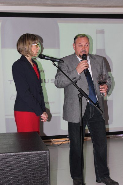 A man and woman speak at a microphone, with a projector screen in the background.