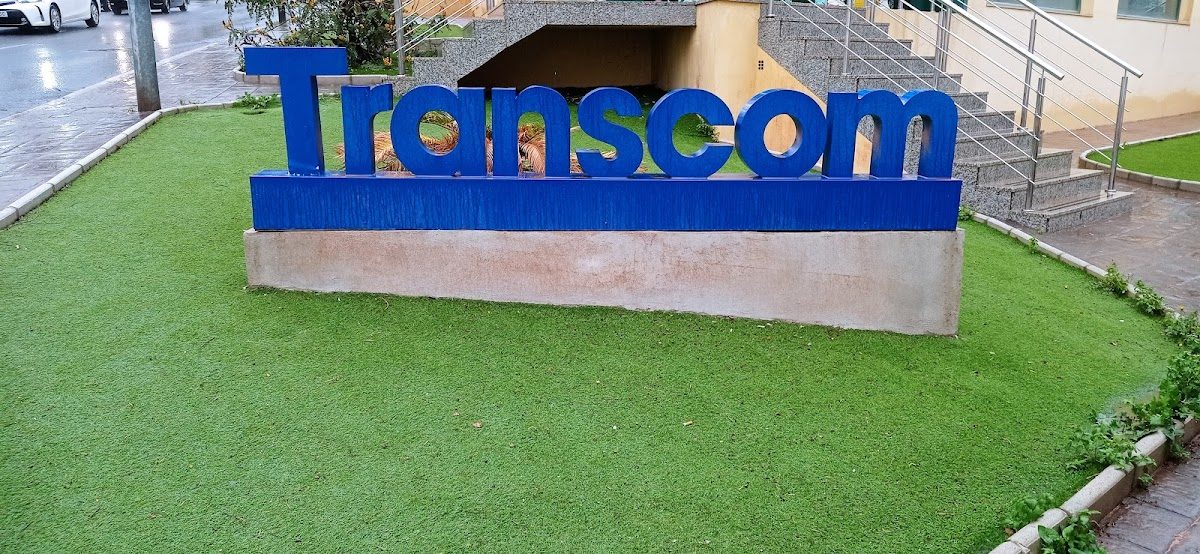 Transcom sign in bold blue letters, set against a green lawn and surrounding pavement.