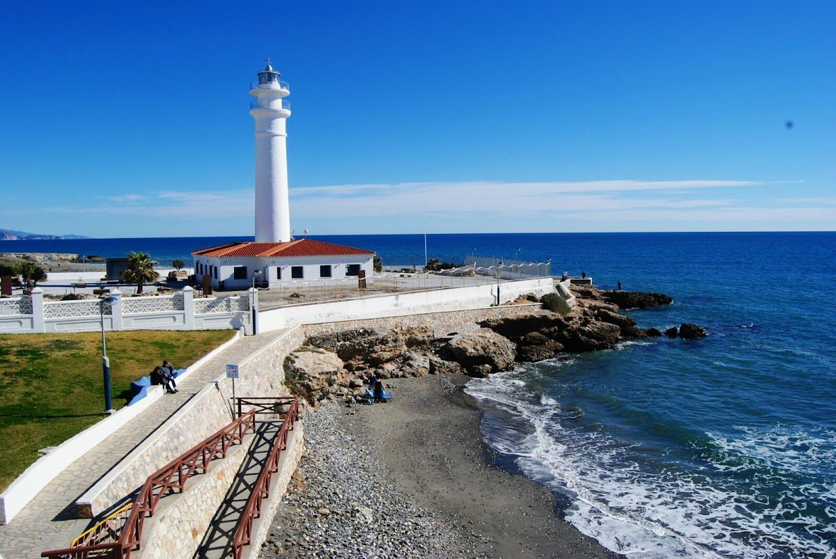The image shows a white lighthouse by the sea, with rocky shores and clear blue skies.