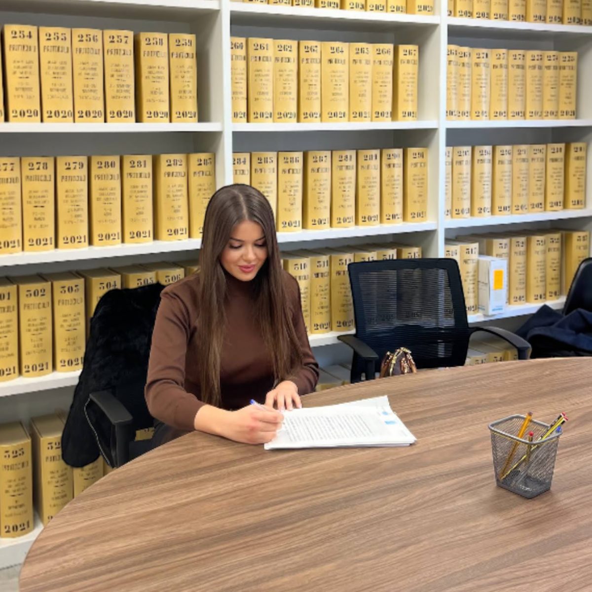 A woman is signing documents at a large wooden table in an office filled with yellow file boxes.