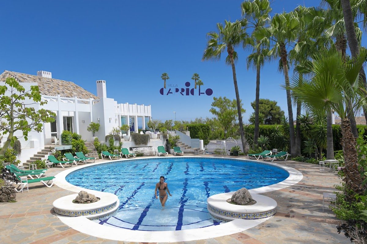 Villa Capricho features a large circular pool surrounded by palm trees and sun loungers in a sunny outdoor setting.
