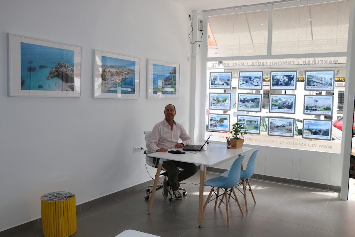 A man sits at a desk in a bright office with framed coastal photos and property listings on the wall.