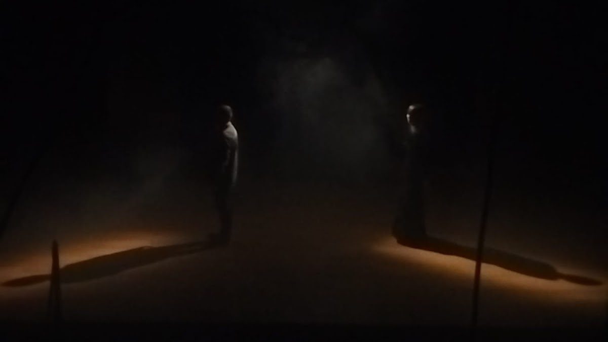 Dos bailarines actúan en el escenario bajo una iluminación tenue, proyectando largas sombras en un ambiente dramático.