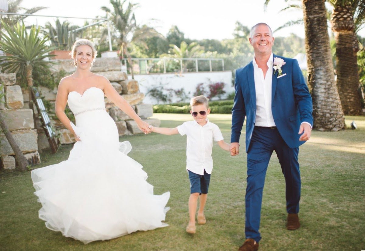 A bride in a white gown walks hand-in-hand with a man in a blue suit and a boy in sunglasses on a green lawn.