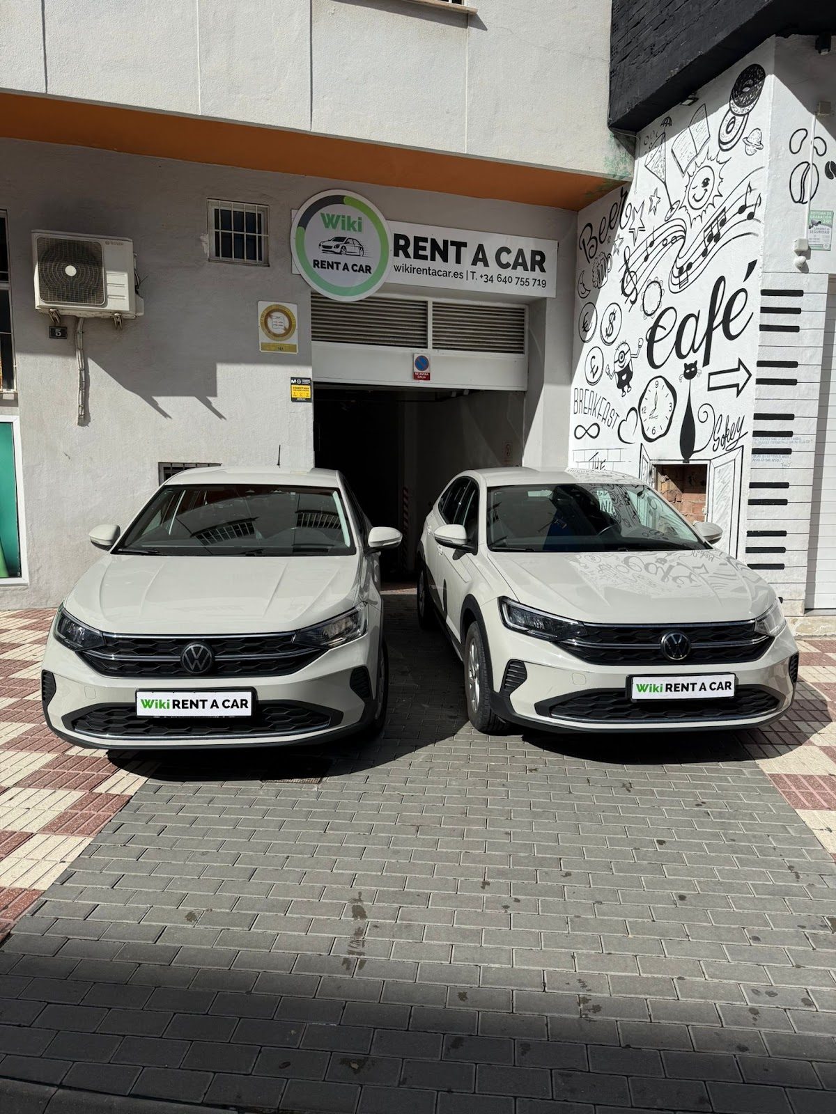 Two white rental cars parked outside Wiki Rent a Car in Estepona, with a mural and signage visible.