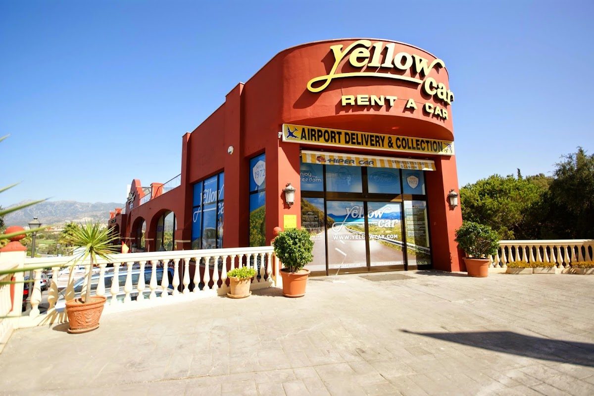 Exterior de color rojo brillante de la agencia de alquiler de coches Yellow Car, con grandes ventanales y plantas en macetas en la entrada.